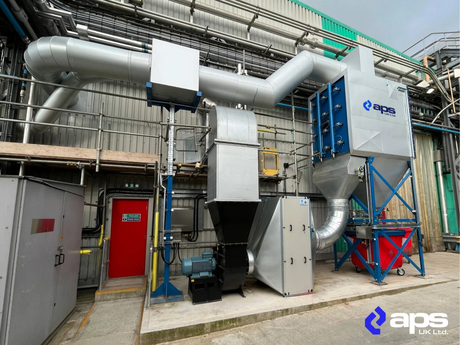 An industrial dust extraction system with large metal ductwork, multiple cylindrical filter housings, and a collection hopper mounted on a blue steel frame, branded with the aps UK Ltd. logo, installed beside a factory building with a red emergency exit door.