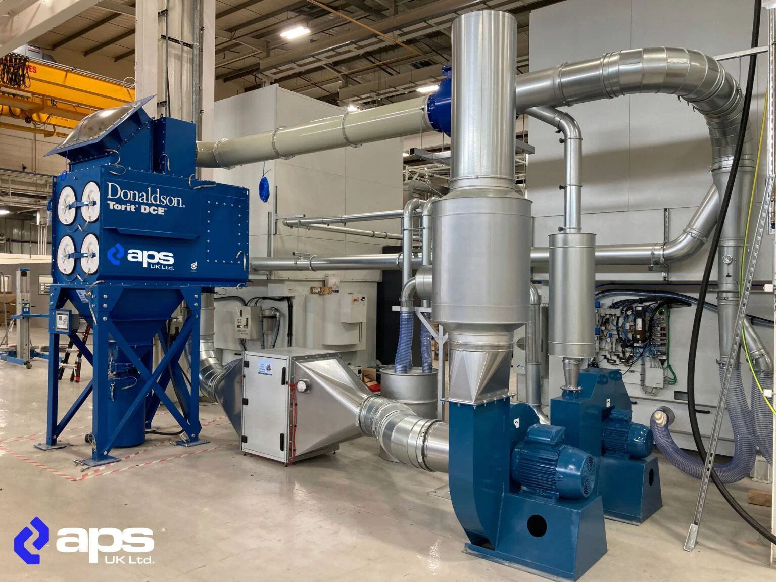 A large blue Donaldson Torit DCE dust extraction unit with multiple circular filter housings and connected metal ductwork, installed inside a manufacturing facility and branded with the aps UK Ltd. logo.