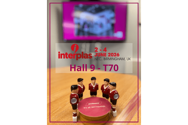 Four tabletop football‑style player figures in red and white stand around a red circular display featuring the Wittmann logo and the phrase ‘It’s all WITTMANN.’ Behind them, an Interplas 2026 graphic shows the event details: 2–4 June 2026, NEC Birmingham, Hall 9 – T70.