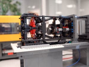 A complex LEGO Technic assembly featuring gears, beams and wiring is mounted on a grey base, resembling a motorised mechanical mechanism, with additional LEGO structures and an indoor office environment blurred in the background.