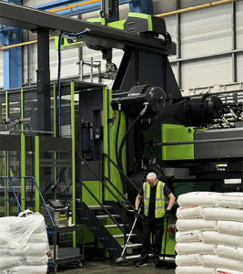 A worker in a high‑visibility vest sweeps the floor beside a large green injection moulding machine with an overhead robotic system and stacked material bags in a modern factory.