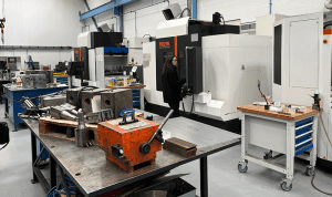 A machinist operates a Mazak CNC machine in a clean, organised workshop with metal components and tools laid out on a central workbench.