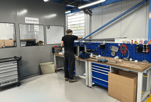 A technician works at a blue‑backed automation workbench with tools, components, and storage drawers in a well‑lit workshop area.