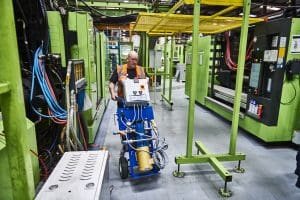 A technician wearing an orange safety vest operates a blue HY‑PRO lifting unit beside large green industrial machinery inside a manufacturing facility.