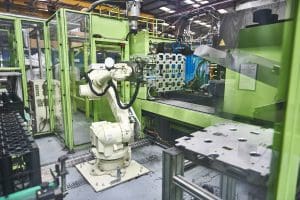 An industrial robotic arm operates next to a large green injection moulding machine, enclosed within a safety cell inside a modern production facility.