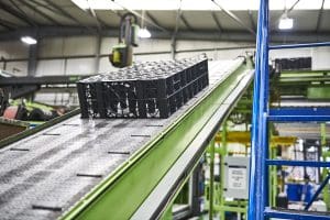 A green automated conveyor system transports stacked black plastic crates upward within a brightly lit, organised manufacturing environment.